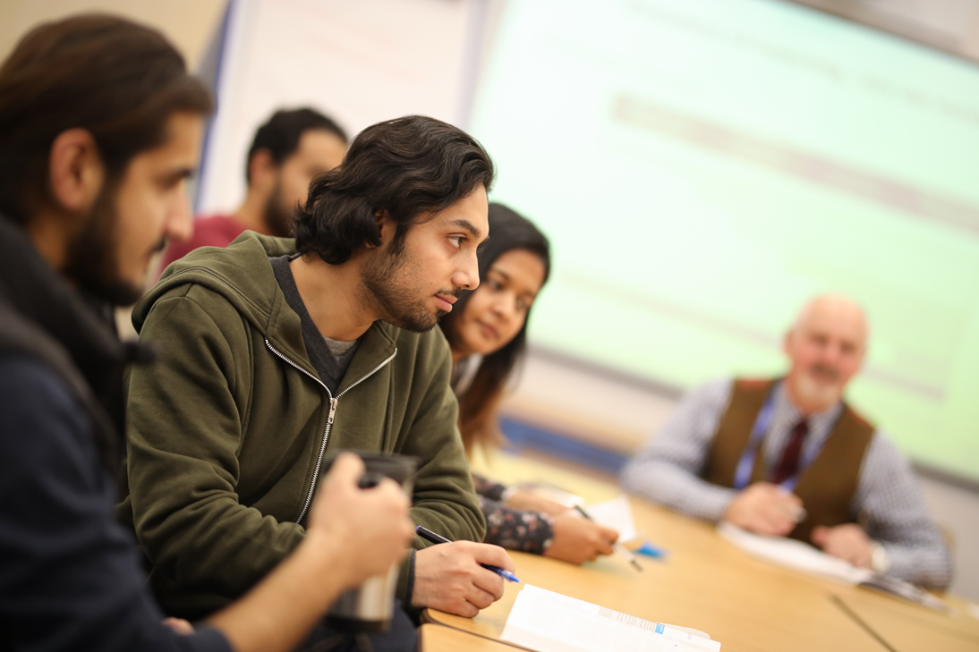 Photograph of a classroom with students in discussion, with a lecturer and projection screen out of focus in the background