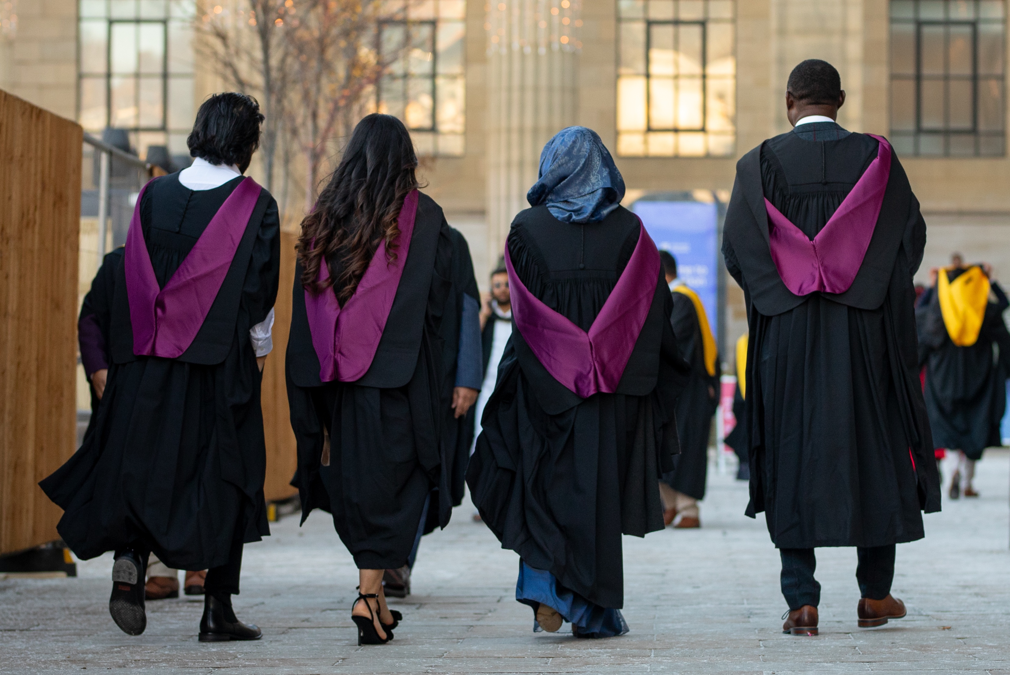 Photograph of four graduands walking away from the camera, wearing graduation robes and hoods. Caird hall and other graduands are blurred in the background.